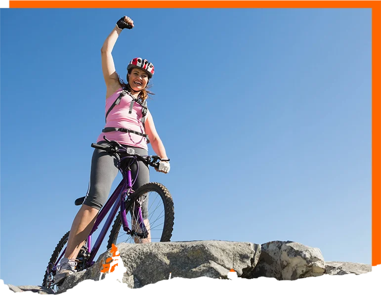 A cyclist stands triumphantly on a rocky outcrop beneath a clear, brilliant blue sky. She’s wearing a helmet and a pink sleeveless top, with one arm raised high in a celebratory fist, giving the scene a strong sense of victory and energy. Her mountain bike is angled on the uneven rock, its thick tires and sturdy frame suggesting a trail or off-road ride rather than a casual street outing. 764 Una ciclista se yergue triunfante sobre un saliente rocoso bajo un cielo azul brillante y despejado. Lleva casco y una camiseta rosa sin mangas, con un brazo en alto en un puño de celebración, lo que confiere a la escena una fuerte sensación de victoria y energía. Su bicicleta de montaña está inclinada sobre la roca irregular; sus neumáticos gruesos y su robusto cuadro sugieren una ruta de montaña o todoterreno, más que un paseo urbano.